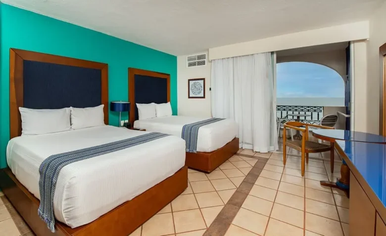 Habitación con dos camas dobles, decoración en tonos azul y turquesa, escritorio y balcón con vista al mar en un entorno tropical.